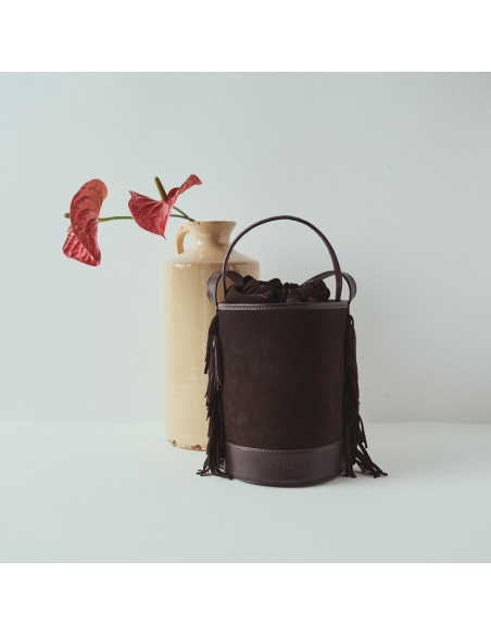 Small brown suede leather bucket bag Roma with fringes, front view, placed in front of a vase with a flower - MONPIEL
