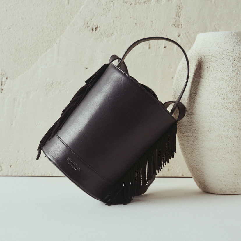Bolsa pequena tipo bucket em couro preto Roma com franjas, vista frontal apoiada sobre um vaso - MONPIEL