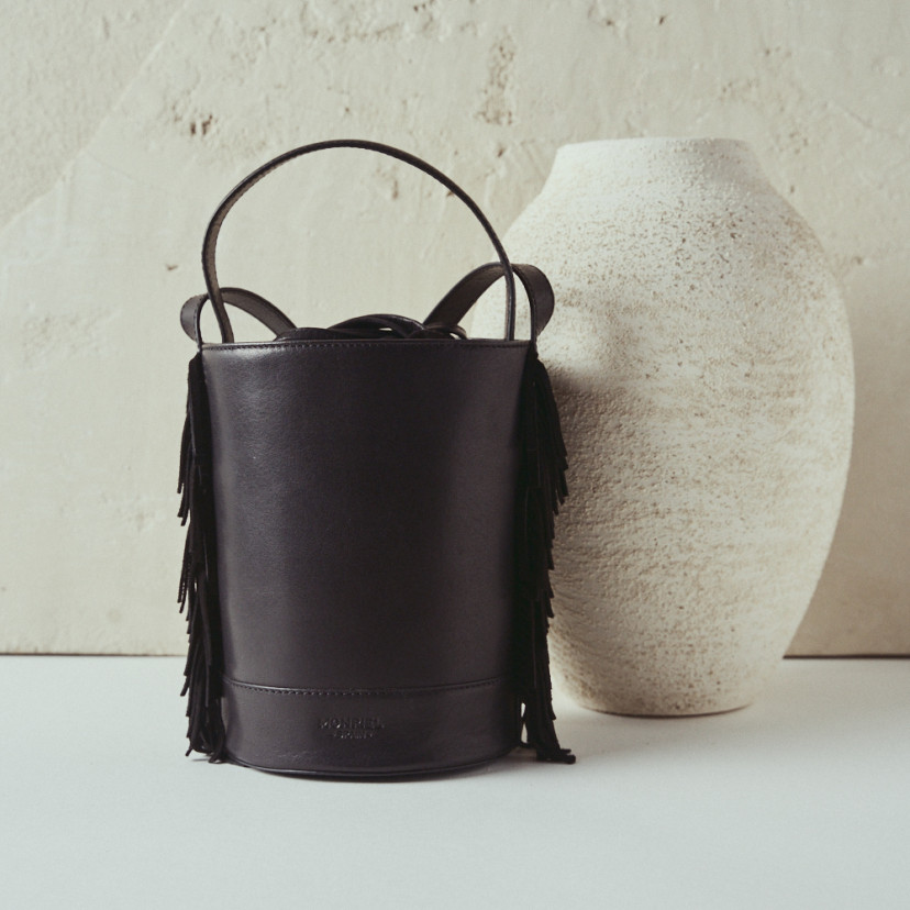 Bolsa pequena tipo bucket em couro preto Roma com franjas, vista frontal ao lado de um vaso - MONPIEL