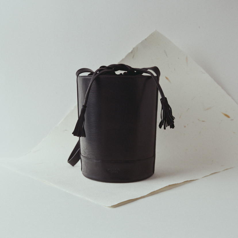 Bolsa redonda em formato de balde de couro na cor preta, vista frontal com detalhe de papel - MONPIEL