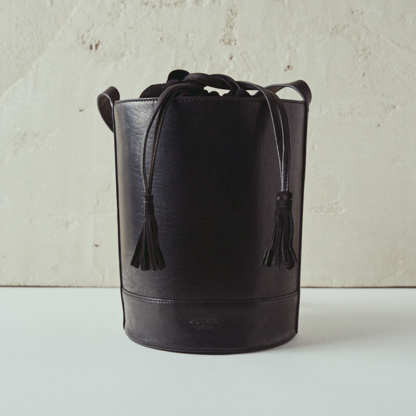 Bolsa redonda em formato de balde de couro na cor preta, vista frontal com fundo de parede - MONPIEL