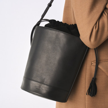 Bolsa redonda em formato de balde de couro na cor preta, vista frontal com fundo de parede - MONPIEL 2