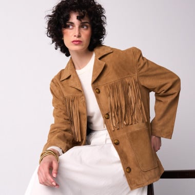 Giacca in pelle scamosciata color cammello con frange, vista in piano generale con modella appoggiata a una poltrona - MONPIEL 2