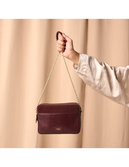 Bolsa tiracolo com corrente dourada combinando pele de serpente e camurça bordô em primeiro plano, segurada pela asa - MONPIEL
