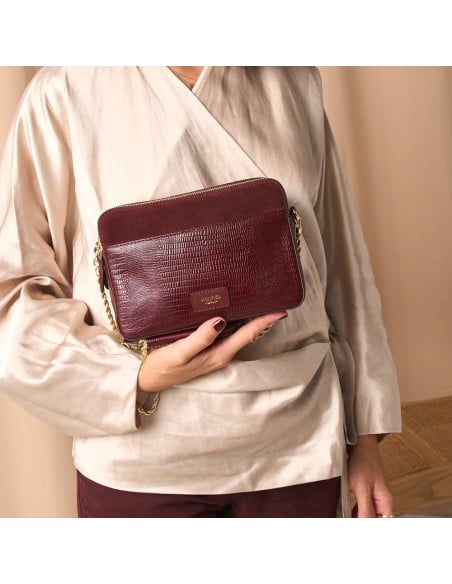 Bolsa tiracolo, corrente dourada combinando pele de serpente e camurça bordô segurada pela base, plano médio da modelo - MONPIEL