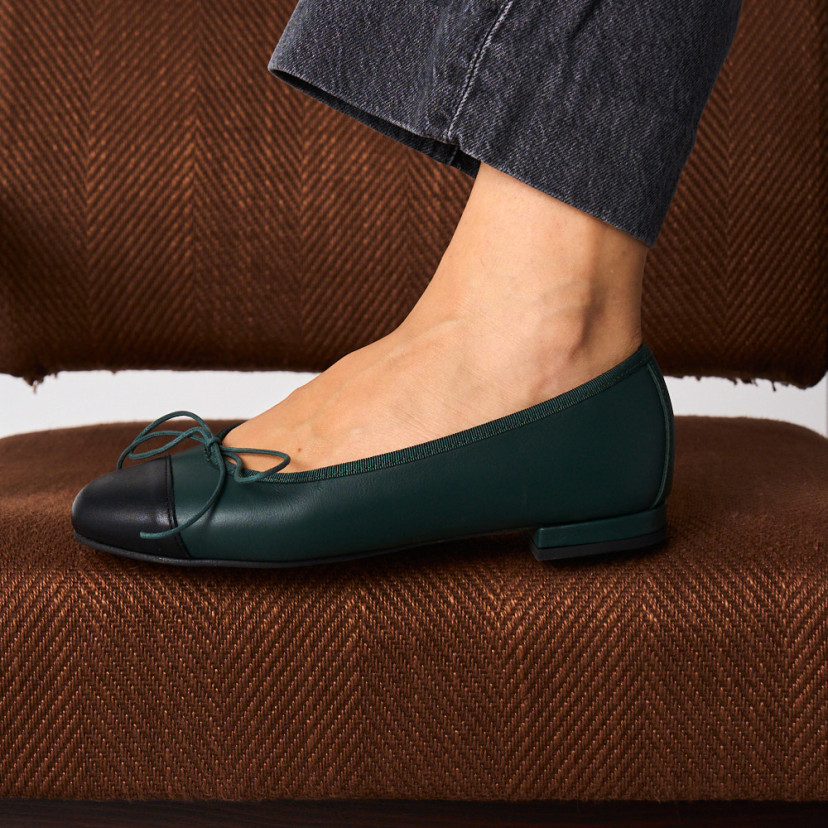 Green leather flat ballerinas with black toe cap, side view of one foot resting on a brown armchair - MONPIEL