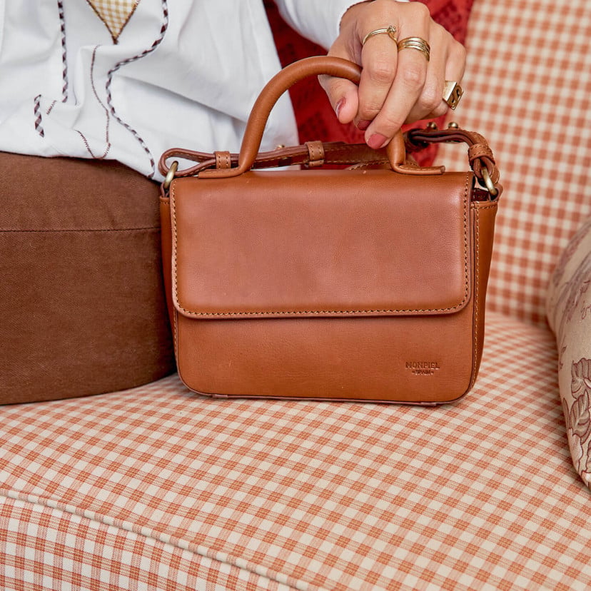 Bolsa pequena de couro Armado em tom couro com vista frontal sobre sofá e alça curta segurada - MONPIEL